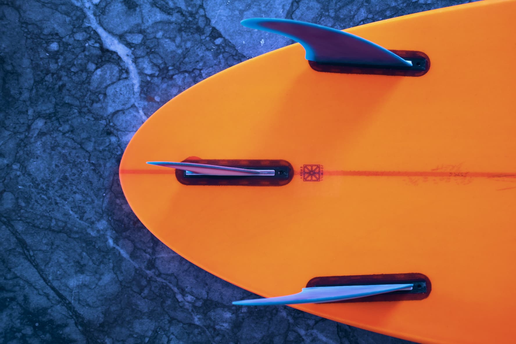 Orange surfboard tail with three blue fins, lying on dark blue stone